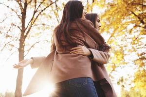 vue depuis l'arrière de jeunes filles jumelles brunes souriantes s'embrassant dans un manteau décontracté se tenant près les unes des autres dans un parc ensoleillé d'automne sur fond flou photo