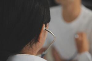 Jeune médecin est en utilisant une stéthoscope Ecoutez à le battement de coeur de le patient. coup de une femelle médecin donnant une Masculin patient une vérifier en haut photo