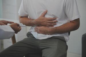 homme Souffrance avec sévère estomac douleur séance à maison. main de mature gars en portant abdomen Souffrance de mal, la diarrhée ou indigestif problème. caucasien gars pressage sur ventre sur douloureux sensation photo