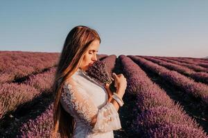 femme lavande champ. content insouciant femme dans une blanc robe en marchant dans une lavande champ et odeur une lavande bouquet sur le coucher du soleil. idéal pour chaud et inspirant concepts dans esprit d'aventure et Voyage. photo