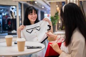 deux asiatique femmes asseoir dans une café à une centre commercial, un avec enthousiasme montrant de une Nouveau chemise à sa ami. photo