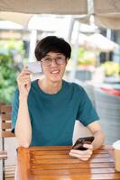 une souriant Jeune asiatique homme montrant le sien crédit carte tandis que séance à une en bois table en plein air. photo