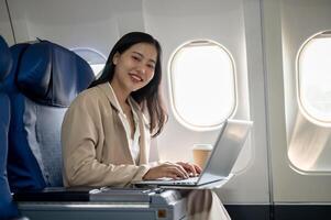 un attrayant asiatique femme d'affaires est sur une vol, séance à une fenêtre siège avec sa portable. photo