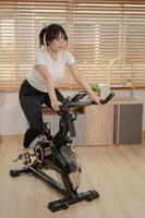 une fort asiatique femme dans tenue de sport est exercice sur une aptitude vélo, Faire intérieur cardio à maison. photo