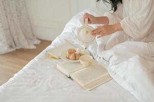 une femme dans longue robe pyjamas verser thé de une magnifique céramique théière, ayant petit déjeuner dans lit photo