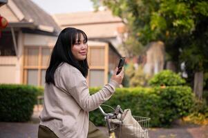 une beau, content Jeune asiatique femme est en utilisant sa téléphone intelligent tandis que équitation une bicyclette dans le ville. photo