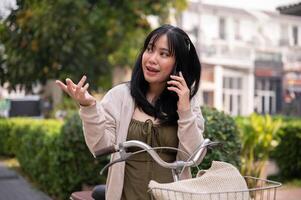 une beau, content Jeune asiatique femme est parlant sur le téléphone tandis que équitation une bicyclette dans le ville. photo