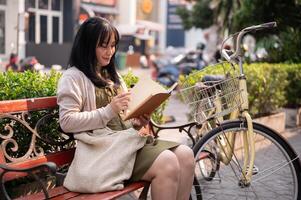 une positif asiatique femme est en train de lire une livre tandis que séance sur une banc en plein air dans le ville avec sa bicyclette photo