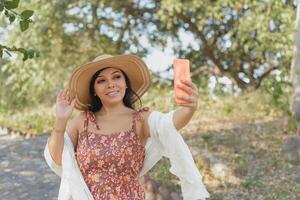 femme portant une chapeau et fleur robe prise une selfie dans une Publique parc. ensoleillé journée. photo