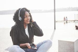 portrait d'une belle jeune jolie fille afro-américaine assise sur la plage ou le lac et écoutant de la musique dans ses écouteurs