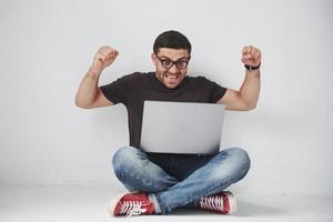 jeune homme caucasien assis sur un mur de briques blanches à l'aide d'un ordinateur portable heureux sous le choc avec un visage surpris, effrayé et excité par l'expression de la peur photo