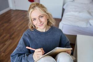 proche en haut portrait de magnifique Jeune femme, l'écriture dans sa agenda, journal ou planificateur, séance dans une chambre, souriant et à la recherche à caméra photo