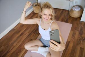 portrait de blond Jeune femme, aptitude entraînement, séance sur yoga tapis, prise selfies pendant faire des exercices à maison, montrant muscles, fléchissement biceps photo