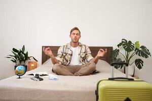 portrait de Jeune conscient voyageur, homme séance sur lit et méditer, emballé valise, Aller à Voyage à l'étranger, entraine toi yoga, a une globe et caméra mensonge suivant à lui photo