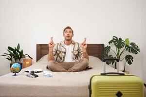 portrait de Jeune homme méditer avec les doigts franchi, prier, fabrication une souhait, emballage valise à aller sur vacances, a globe et caméra sur une lit photo