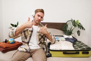 portrait de de bonne humeur Beau homme, touristique Aller sur vacances, dansant et écoute à la musique dans écouteurs tandis que emballage valise avec vêtements pour vacances voyage à l'étranger photo