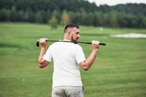 tenant le bâton derrière. portrait de joueur de golf marchant dans la pelouse et équipement en main. bois à l'arrière-plan photo