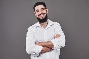 jeune bel homme appuyé contre un mur gris avec les bras croisés. un jeune homme sérieux avec une barbe regarde la caméra photo