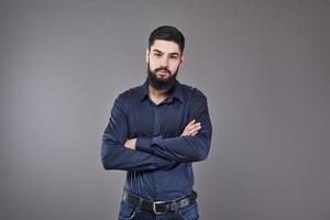 jeune bel homme appuyé contre un mur gris avec les bras croisés. un jeune homme sérieux avec une barbe regarde la caméra photo