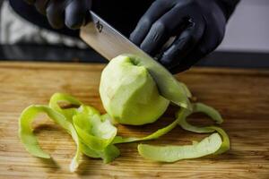 la personne avec noir gants peeling et Coupe un Pomme avec Japonais couteau photo