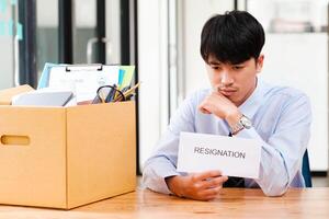 homme à bureau avec démission lettre, méditer le sien décision à côté de une emballé boîte. photo