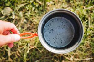 l'eau dans le pot à le ébullition organiser, bulles à le bas de le poêle, main en portant le vaisselle par le poignées, camp ustensiles, une tasse pour cuisson. photo