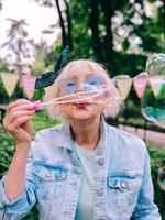 femme élégante senior aux cheveux gris et à lunettes bleues et veste en jean soufflant des bulles à l'extérieur. vacances, fête, anti-âge, concept amusant photo
