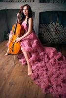 Portrait de belle jeune femme aux longs cheveux noirs avec violoncelle en longue robe rose en studio photo