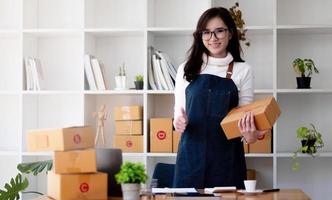 heureuse jeune femme asiatique démarrage petite entreprise indépendante tenant une boîte à colis et un ordinateur portable et assise sur une chaise, concept de livraison de boîte d'emballage marketing en ligne photo