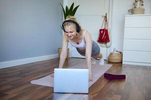 portrait de Jeune sportif femme Faire entraînement, à la recherche à aptitude sur portable dans sans fil écouteurs, permanent dans planche sur yoga tapis, Suivant exercice Didacticiel photo