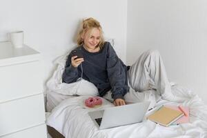 mode de vie et fin de semaine concept. Jeune souriant femme, relaxant à maison, mensonge dans lit avec tasse de thé et Donut, en utilisant portable et téléphone intelligent dans chambre photo