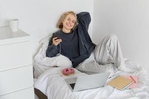 portrait de Jeune magnifique blond femme, sentiment paresseux à maison, mensonge dans lit avec tasse de thé, Donut et ordinateur portable, en train d'étudier à distance ou en train de regarder film en ligne photo