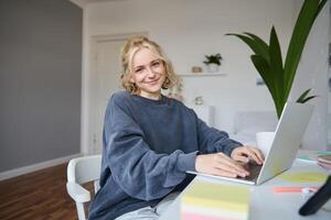 portrait de Jeune souriant magnifique femme, étudiant en train d'étudier à distance dans sa chambre, e-learning, en utilisant ordinateur portable, séance sur chaise dans chambre à coucher, à la recherche content à caméra photo