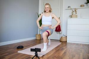 portrait de femme, aptitude instructeur à maison, enregistrement à propos entraînement, montrant Comment à faire jambe des exercices, squats avec élastique la résistance groupe, travail en dehors à l'intérieur sur yoga tapis photo