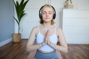 portrait de Jeune détendu femme, est assis dans pièce dans écouteurs, fermoir mains ensemble, médité, écoute à yoga la musique photo