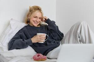 portrait de Candide, content Jeune femme mensonge dans lit, à la recherche à portable filtrer, en portant tasse de thé et en mangeant Donut, rester à Accueil sur fin de semaine, dépenses assez temps seul photo