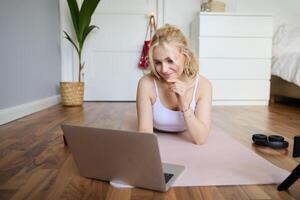 portrait de magnifique blond femme à la recherche à aptitude tutoriels sur ordinateur portable, mensonge sur caoutchouc yoga tapis, Suivant faire des exercices instructions en ligne photo