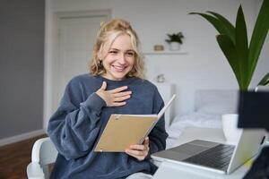 portrait de souriant Jeune mignonne femme, en portant carnet de notes, fille avec portable et planificateur dans mains, est assis dans chambre, regards à numérique caméra, crée mode de vie vlog, pourparlers à public, lit scénario photo