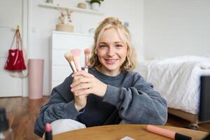 portrait de Jeune femme, contenu créateur, fabrication une à propos se maquiller, montrant brosses à public, à la recherche à caméra, enregistrement beauté Didacticiel, souriant Heureusement photo