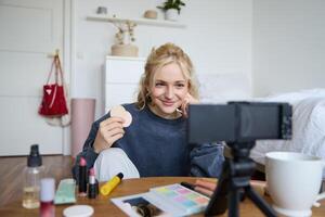 image de Jeune femme, vlogger enregistrements sur numérique caméra, spectacles beauté des produits, recommander maquillage pour public sur social médias, est assis sur de face dans une pièce photo