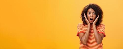 à la taille coup de sous le choc concerné paniquer Afro-américain femme avec afro coiffure dans branché T-shirt halètement en portant paumes sur visage de surprise posant troublé contre Orange mur photo