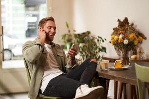 mode de vie coup de Beau Jeune homme, à la recherche à le sien mobile téléphone, écoute à la musique dans sans fil écouteurs, séance dans une café, en buvant café photo