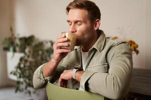 café, loisir et mode de vie concept. portrait de Beau Jeune homme est assis sur chaise avec verre de café, les boissons le sien boisson et souriant photo