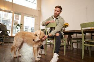 proche en haut portrait de Beau Jeune homme attendre pour le sien commande dans café boutique, caresse une chien, repos dans acceptant les animaux café photo