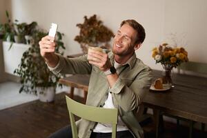 portrait de Jeune Beau homme prend slefie sur le sien mobile téléphone application, est assis dans café avec verre de café, affectation sur social médias application photo