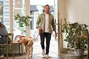portrait de Beau Jeune homme des promenades dans le café avec le sien chien sur une laisse, entre dans acceptant les animaux café boutique, ouvre le porte photo