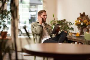 portrait de Beau content gars, Jeune homme est assis dans café, en train de regarder s sur numérique tablette, portant sans fil écouteurs, en riant et souriant, dépenses temps dans café magasin photo