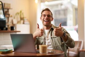 portrait de Beau souriant homme, spectacles les pouces en haut, est assis dans café avec tasse de café et ordinateur portable, approuve qch bien, recommande cotravail espace ou en ligne site Internet photo