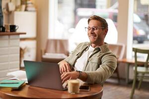 portrait de homme travail dans café sur ordinateur portable, à la recherche satisfait et heureux avec lui-même, portant des lunettes pour ordinateur écrans, est assis retour sur chaise dans café magasin photo