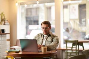 image de Jeune numérique nomade, homme dans des lunettes est assis dans café, travaux de café boutique, les usages portable dans cotravail espace, porte lunettes, les boissons le sien boisson photo